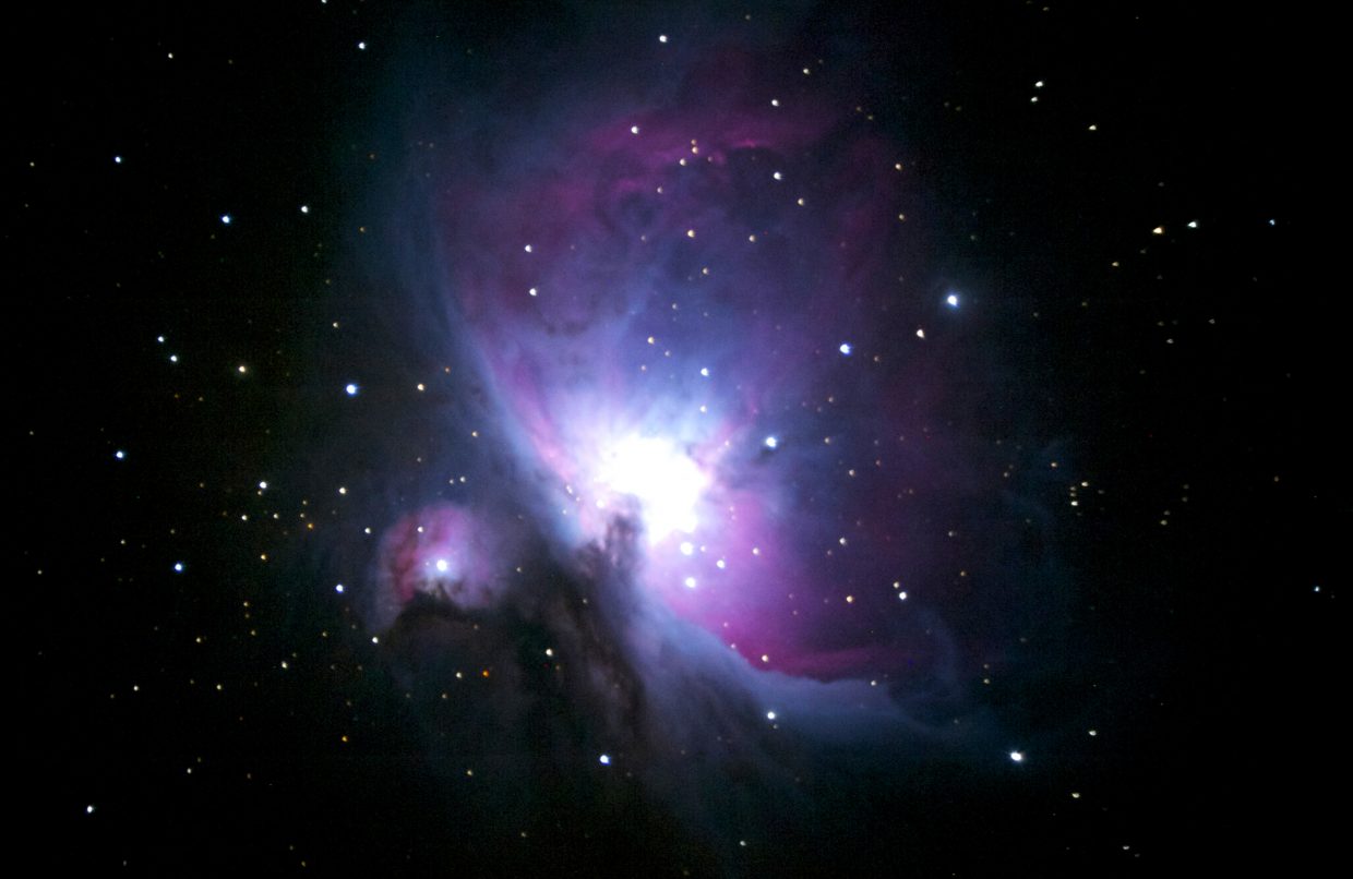 a photograph of the orion nebula. it is a purple and blue gaseous structure with a bright white center. it is surounded by the black expanse of space and some distant stars